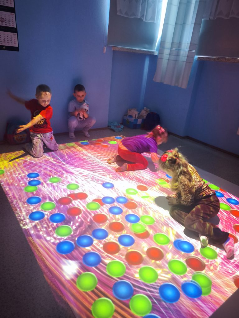 Cztery dzieci w jasnym pokoju na podłodze z kolorowymi, okrągłymi wzorami świetlnymi, ściany i zasłony w odcieniach niebieskiego.