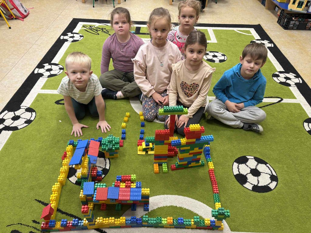 Sześć dzieci siedzi na zielonym dywanie z motywem piłek nożnych, przed nimi różnokolorowa budowla z klocków Lego.