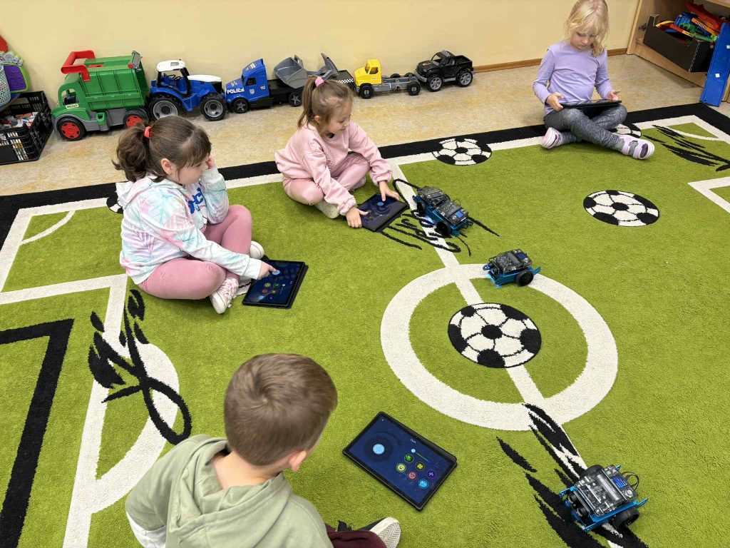 Cztery dzieci siedzące na zielonym dywanie z motywem boiska piłkarskiego, każda z tabletem, obok trzy małe roboty, w tle zabawkowe pojazdy.