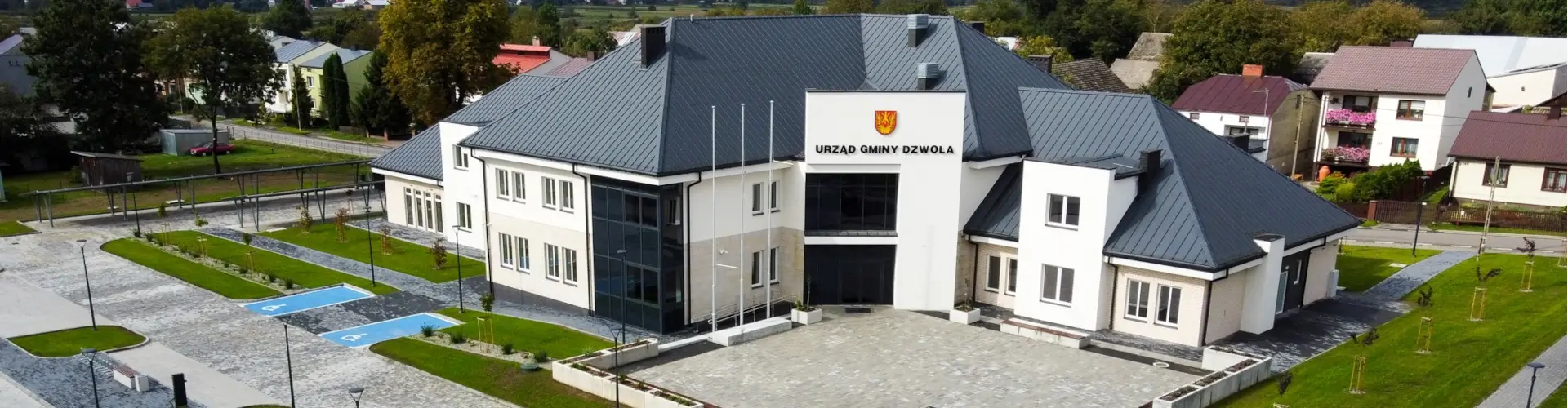 Biały budynek z ciemnoszarym dachem, szklany narożnik, przed wejściem trzy maszty flagowe, wokół zielone trawniki i chodniki, dwa niebieskie miejsca parkingowe dla niepełnosprawnych.