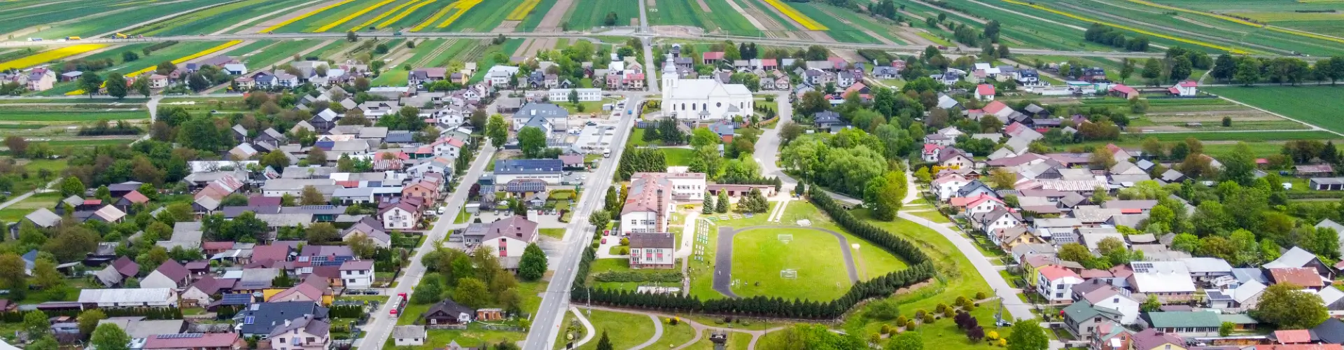 Mała miejscowość z białym kościołem, domami o różnych dachach, zielonym boiskiem otoczonym drzewami, polami uprawnymi w tle.