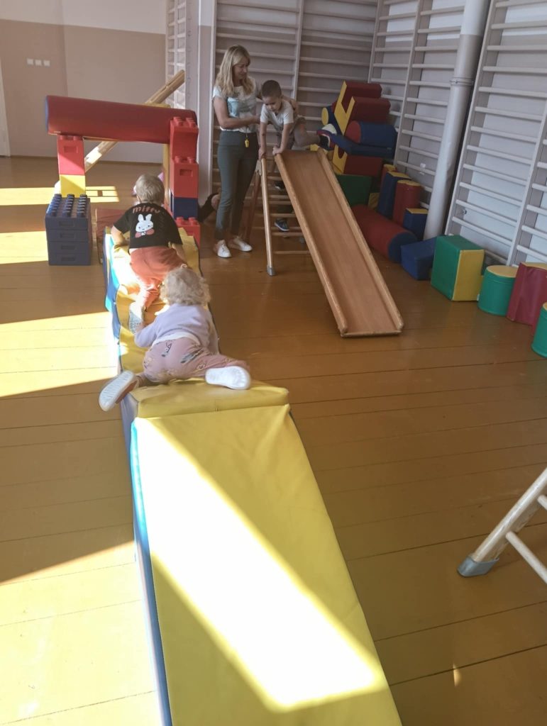 Dzieci bawiące się na miękkich kolorowych materacach i drewnianej ślizgawce w jasnym pomieszczeniu z drewnianą podłogą, obok stoi kobieta.