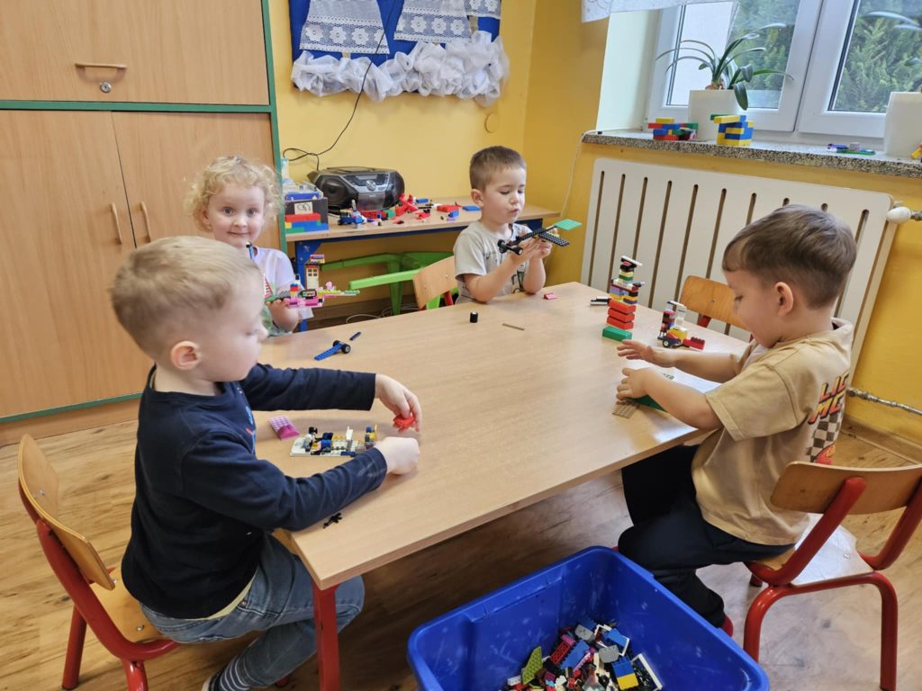 Cztery dzieci siedzą przy drewnianym stole w żółtym pokoju, bawią się kolorowymi klockami konstrukcyjnymi, na stole i w niebieskim pojemniku obok stołu.