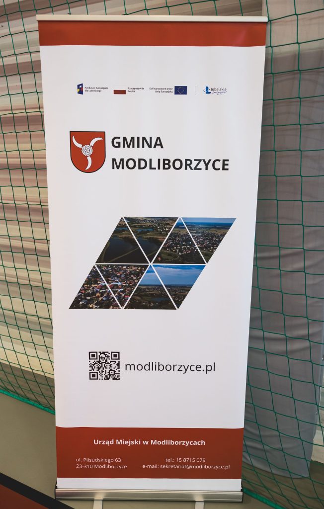 Rollup z herbem i napisem Gmina Modliborzyce, zdjęcia lotnicze ułożone w sześć trójkątów, adres strony modliborzyce.pl, dane kontaktowe Urzędu Miejskiego na czerwonym tle.