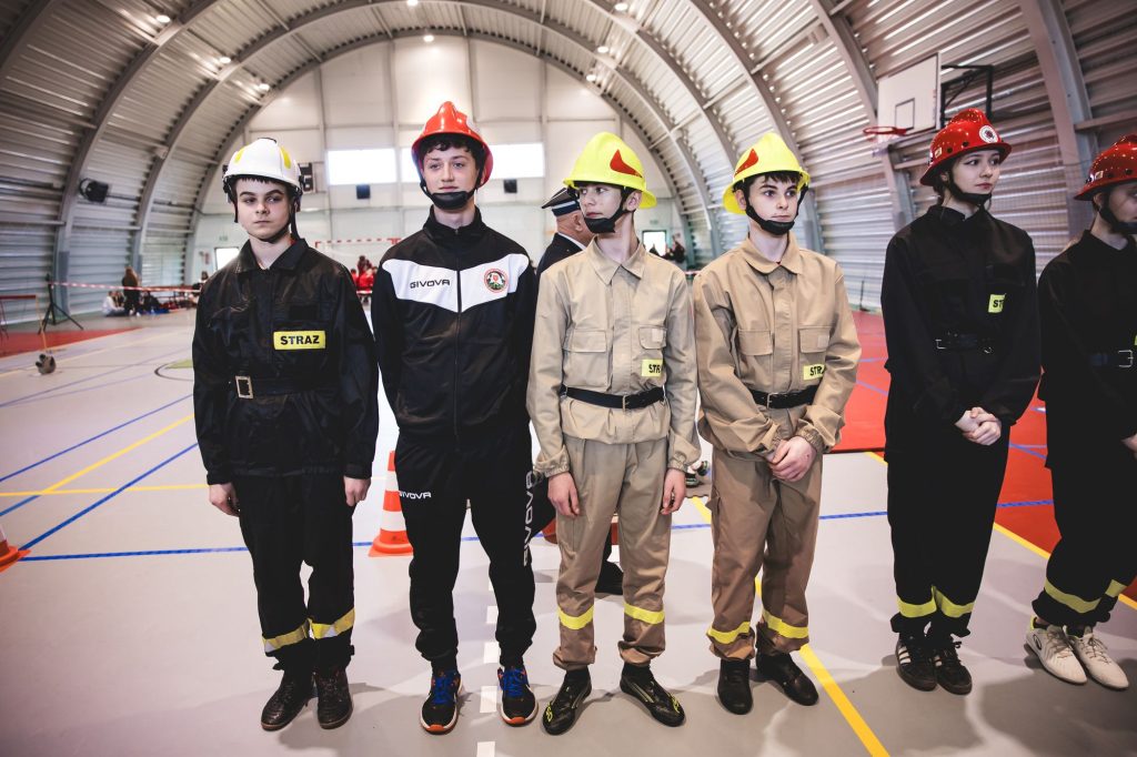 Grupa młodych osób w stroju strażackim i kaskach w hali sportowej na jasnej podłodze z oznaczeniami boiska.