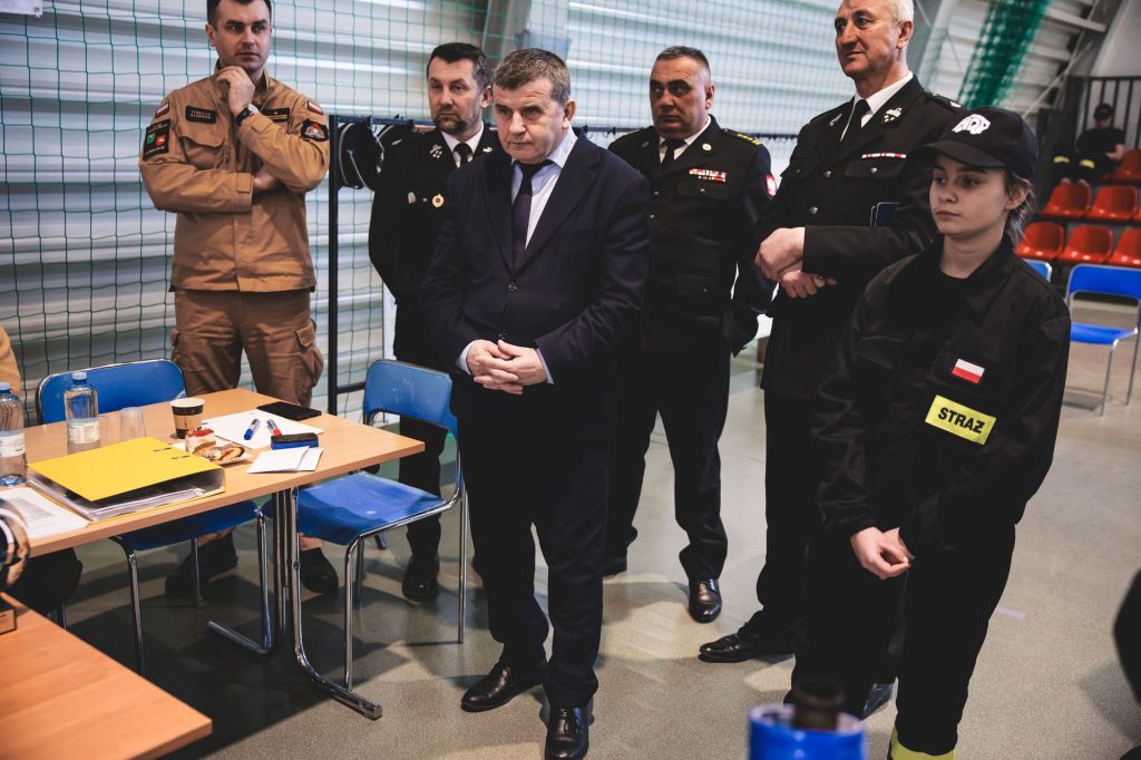 Grupa sześciu osób w mundurach strażackich i garniturze stoi przy stole z dokumentami i napojami, w tle zielona siatka i czerwone krzesła.