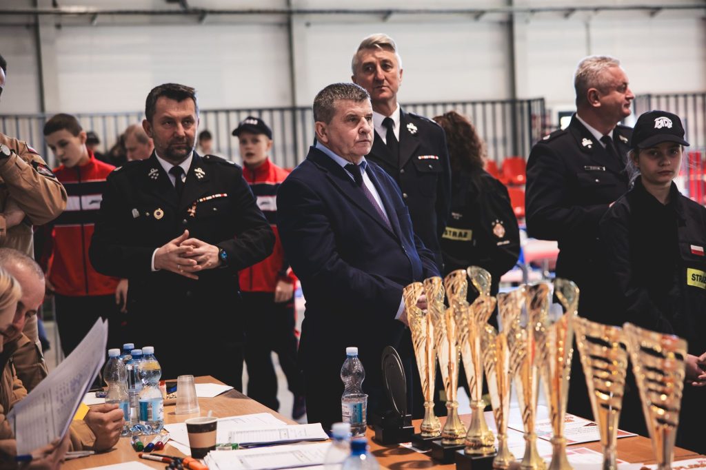 Mężczyźni i kobiety w mundurach strażackich stoją przy stole z dokumentami, butelkami wody i złotymi pucharami w hali sportowej.