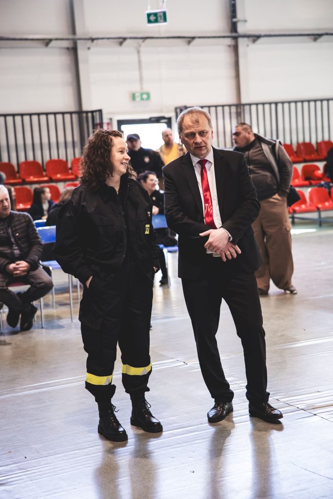 Kobieta w czarnym kombinezonie z żółtymi odblaskami i mężczyzna w czarnym garniturze z czerwonym krawatem stoją na jasnej podłodze w hali z czerwonymi i niebieskimi krzesłami.