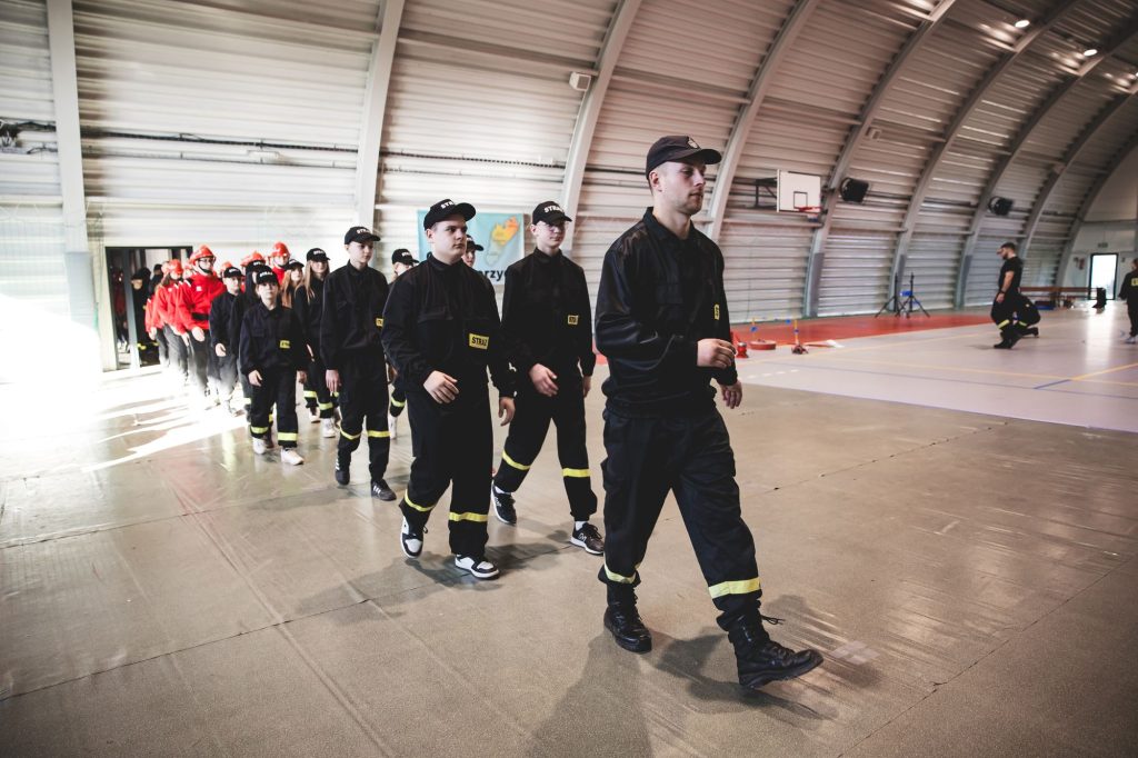 Grupa osób w czarnych mundurach z żółtymi pasami i czapkach wchodzących do hali sportowej, w tle kolejna grupa w czerwonych strojach.