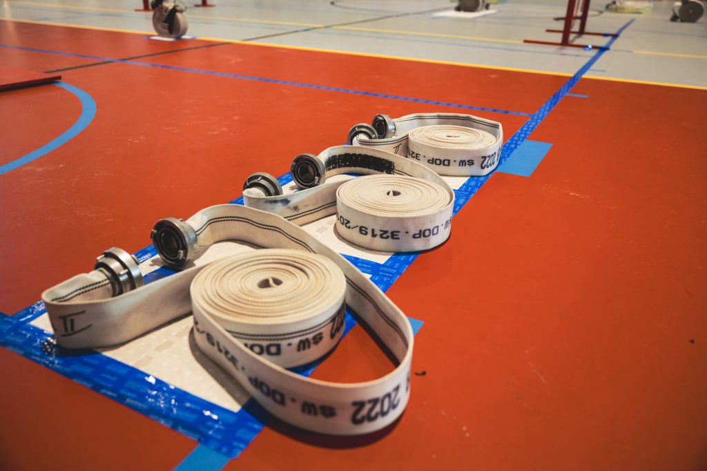 Trzy zwinięte białe węże strażackie z metalowymi zakończeniami na czerwonej podłodze hali sportowej z niebieskimi liniami.