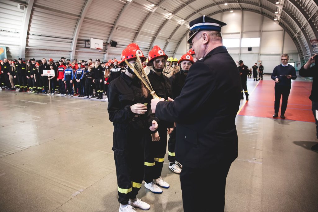 Grupa młodych osób w czarnych kombinezonach i czerwonych hełmach, otrzymujących złoty puchar od mężczyzny w mundurze w hali sportowej.