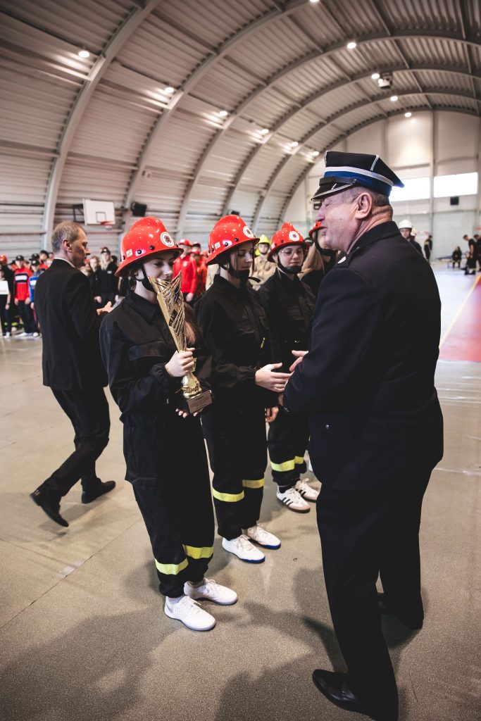Grupa młodych strażaków w czarnych mundurach z żółtymi pasami i czerwonych hełmach stoi w hali, jedna osoba trzyma złoty puchar, obok mężczyzna w mundurze oficerskim.