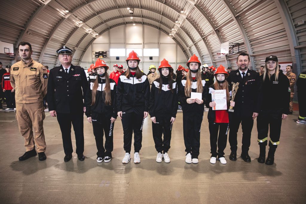 Grupa ludzi w hali sportowej, z przodu pięć dziewczyn w czarnych dresach i czerwonych hełmach, dwie osoby w mundurach strażackich, jedna w beżowym stroju, jedna w czarnym ubraniu.