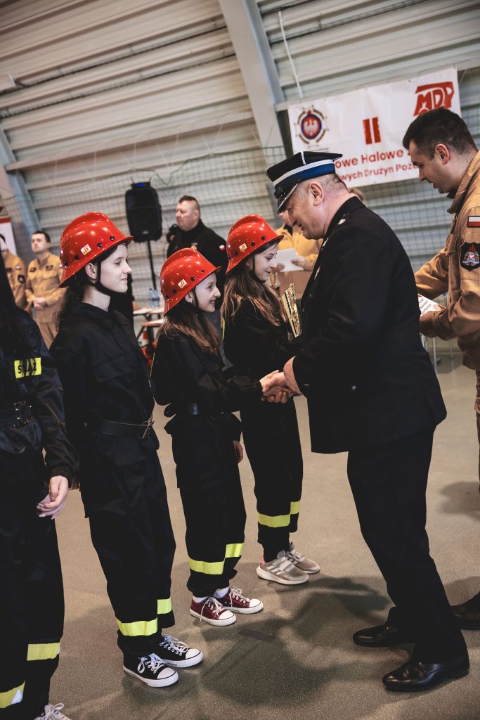 Trzy dziewczynki w czarnych uniformach z żółtymi paskami i czerwonych hełmach, ściskające dłoń mężczyźnie w mundurze, w hali z siatką i banerem w tle.