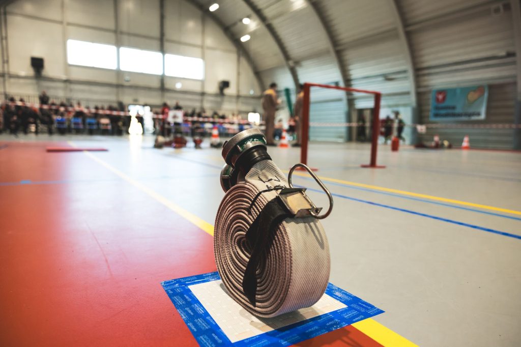 Zwinięty biały wąż strażacki z metalowymi końcówkami na podłodze hali sportowej z czerwoną i szarą nawierzchnią. W tle rozmyci ludzie i sprzęt.