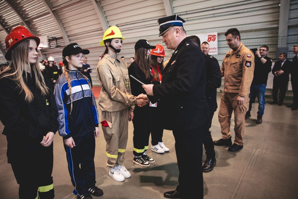 Mężczyzna w mundurze wręcza dokument młodzieży w strojach strażackich i sportowych, na hali z łukowym dachem, w tle kilka osób obserwujących wydarzenie.