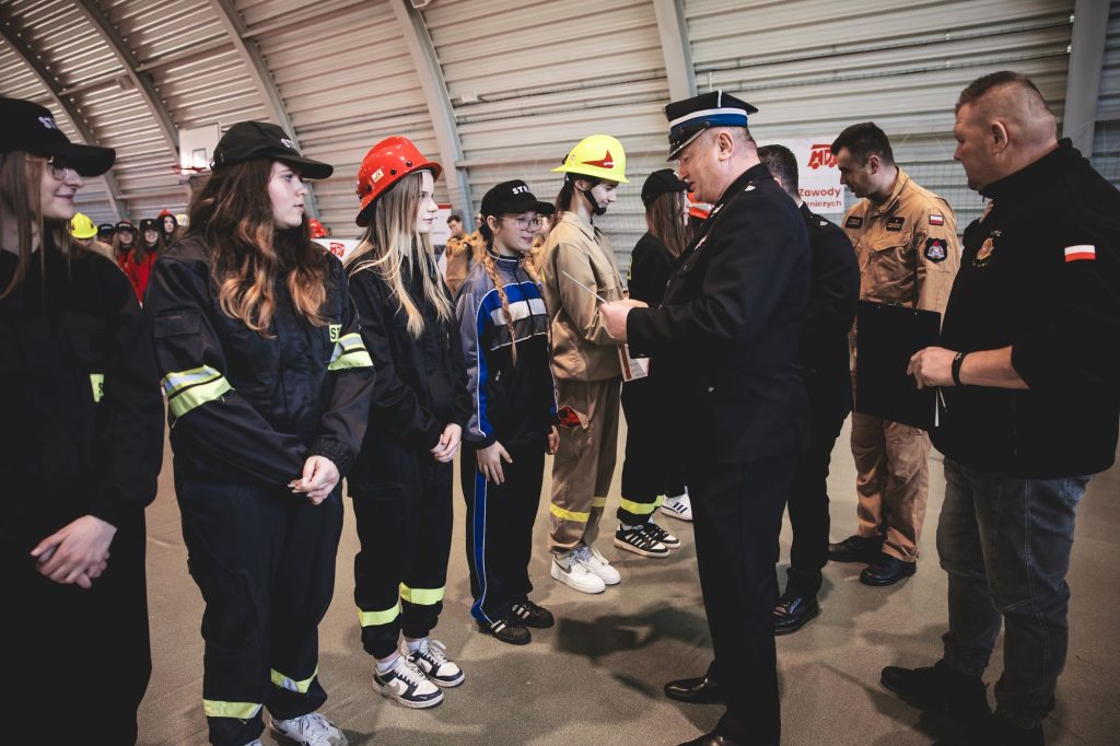 Grupa młodych osób w mundurach strażackich i hełmach stoi w hali, mężczyzna w mundurze wręcza im dokumenty, w tle inni mężczyźni w mundurach i ubraniach roboczych.