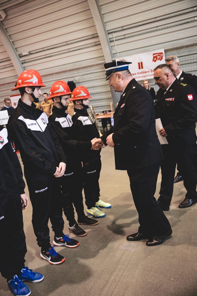 Mężczyzna w czarnym mundurze z granatową czapką podaje rękę chłopcu w czerwym kasku i czarnym dresie, w tle inni chłopcy w podobnych strojach oraz mężczyźni w mundurach.