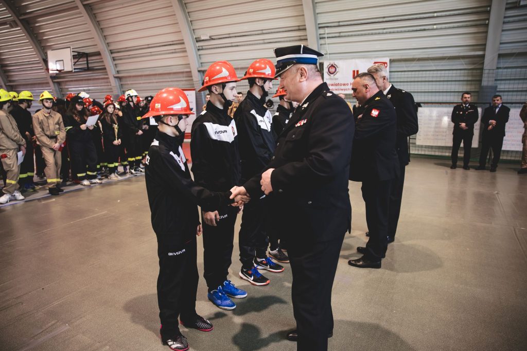 Mężczyzna w czarnym mundurze podaje rękę chłopcu w czerwonym kasku i czarnym stroju sportowym, w tle grupa osób w hełmach i mundurach na hali sportowej.