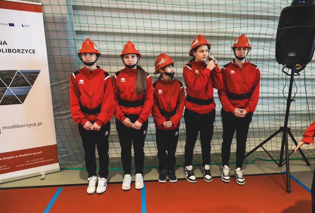Pięć osób w czerwonych kurtkach, czarnych spodniach i czerwonych hełmach stojących na hali sportowej przy siatce i banerze.