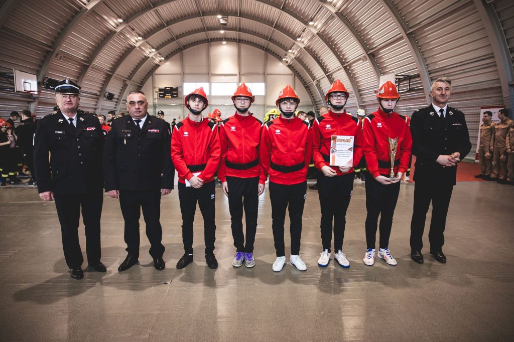 Sześciu chłopców w czerwonych kurtkach, czarnych spodniach i czerwonych hełmach oraz czterech mężczyzn w czarnych mundurach stoi w hali sportowej o metalowym sklepieniu.