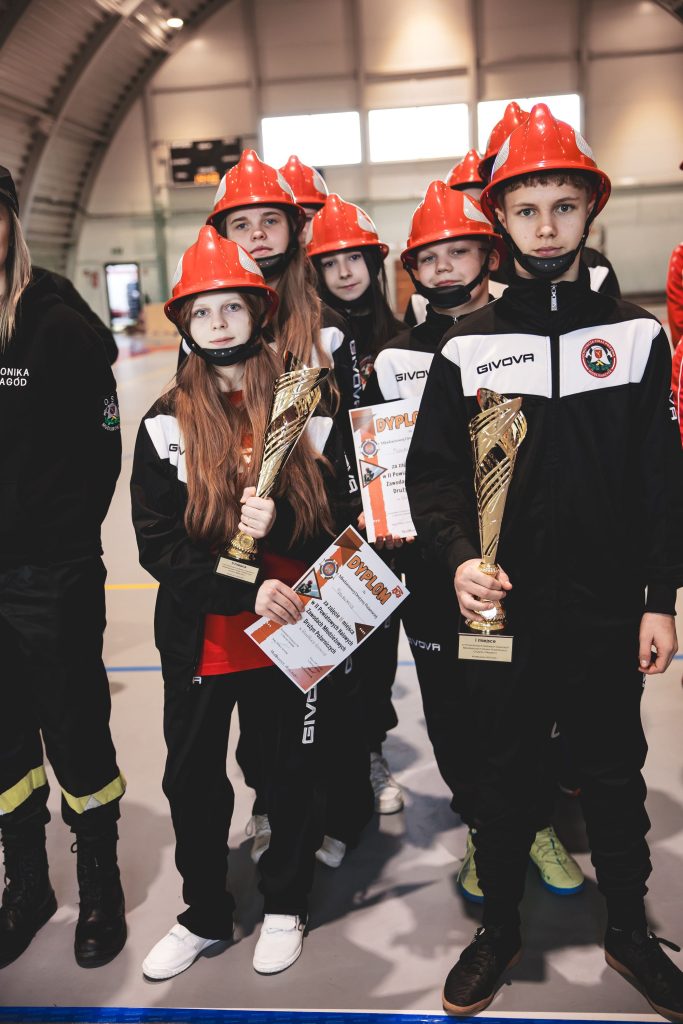 Grupa dzieci w czarno-białych dresach i czerwonych kaskach, trzymająca złote puchary i dyplomy, stojąca na hali sportowej o jasnych ścianach i podłodze.