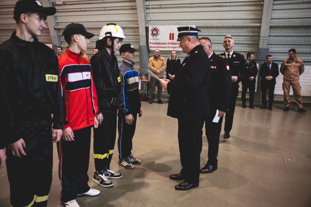 Młodzież w mundurach strażackich stoi w rzędzie naprzeciw mundurowych mężczyzn, hala sportowa, tablica z napisem w tle.
