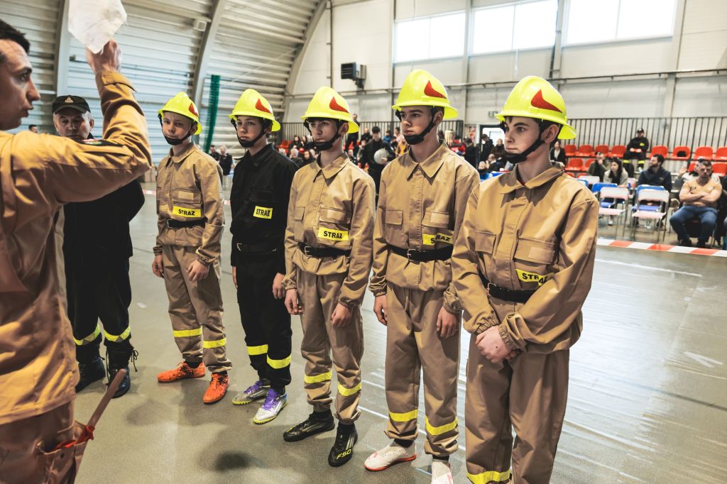 Młodzi chłopcy w beżowych i czarnych mundurach strażackich z żółtymi hełmami stoją w rzędzie w hali sportowej, obok nich mężczyźni w mundurach.
