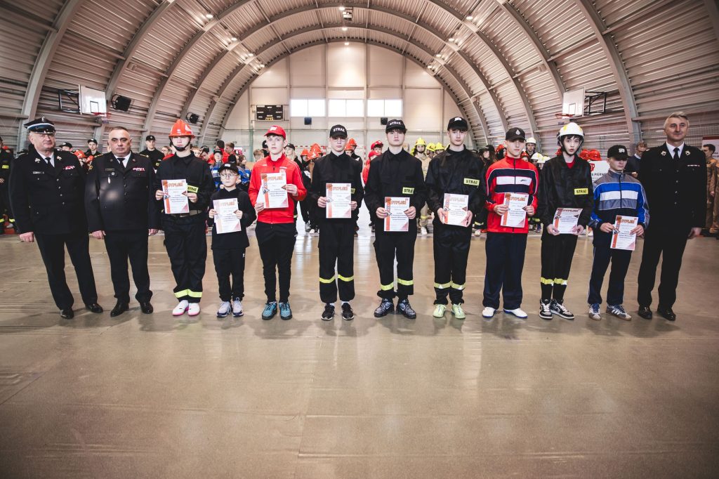 Grupa młodych strażaków w czarnych mundurach i czerwonych czapkach stoi w hali sportowej z dyplomami, obok nich mężczyźni w mundurach oficerskich.