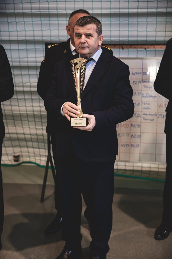 Mężczyzna w ciemnym garniturze trzyma złoty puchar, w tle siatka i tablica z notatkami.