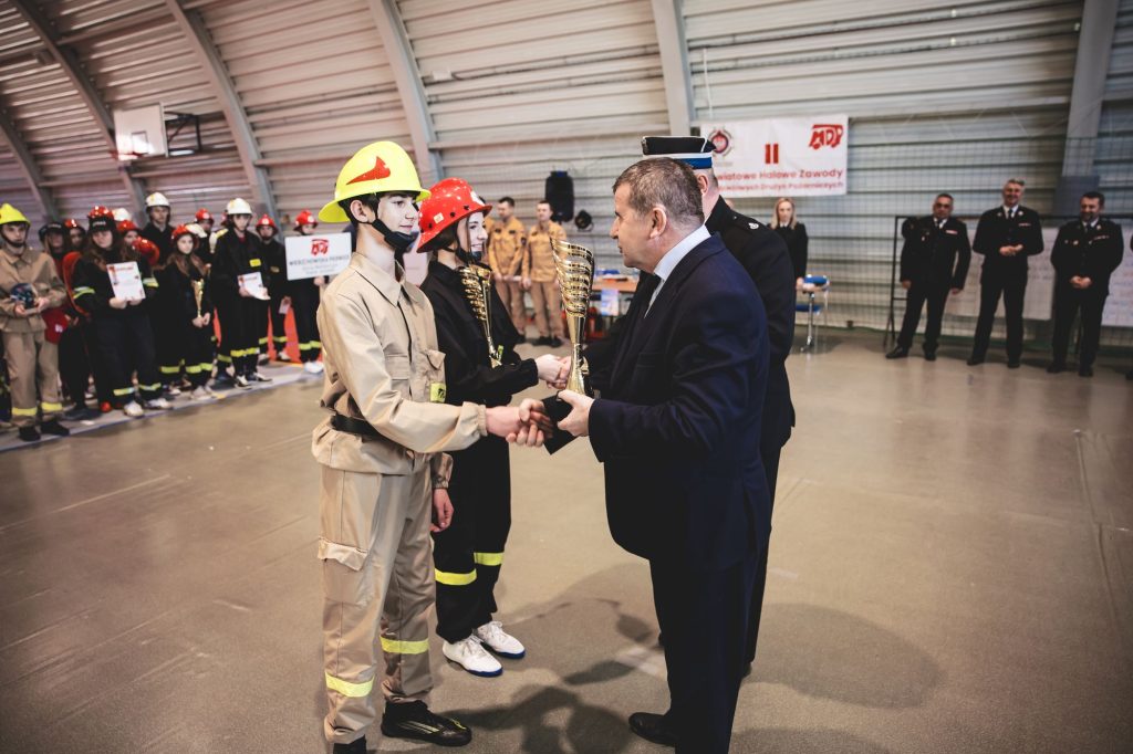Młodzi strażacy w kaskach żółtym i czerwonym odbierają puchary, w tle grupa osób w strojach strażackich i mężczyźni w mundurach na hali sportowej.