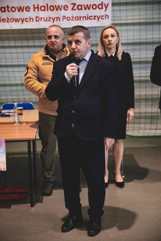 Mężczyzna w ciemnym garniturze trzyma mikrofon, za nim stoi kobieta w mundurze i mężczyzna w beżowej bluzie z napisem OSP, w tle siatka i fragment baneru z czerwonym napisem.