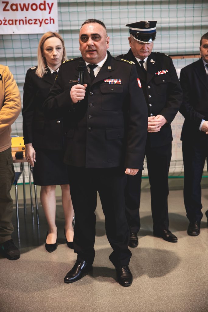 Mężczyzna w czarnym mundurze z odznakami trzyma mikrofon, obok stoi kobieta w czarnym mundurze i mężczyzna w czarnym mundurze z czapką, w tle siatka i fragment napisu czerwonym drukiem.