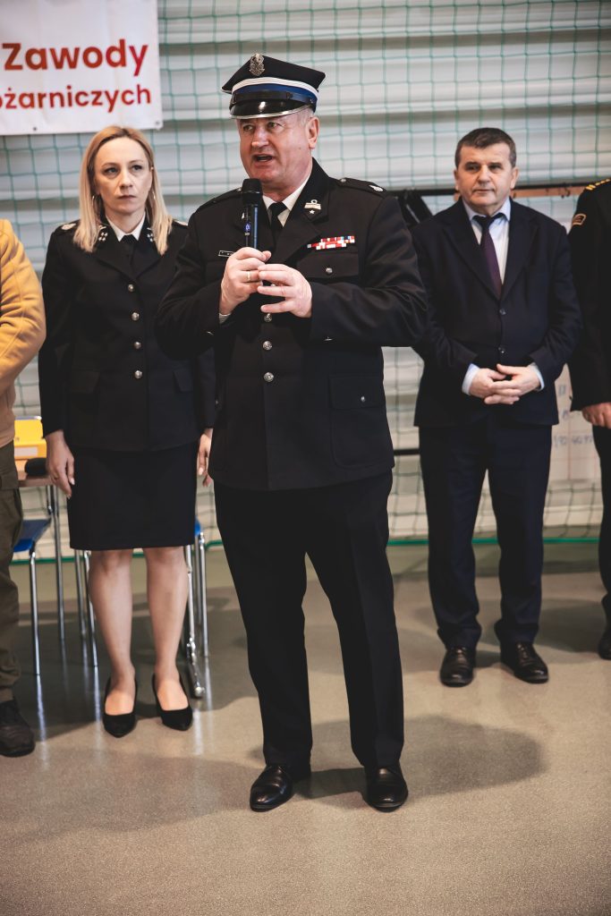 Mężczyzna w mundurze strażaka z mikrofonem, obok kobieta w mundurze i mężczyzna w garniturze, w tle siatka zielona i fragment tablicy z napisem.