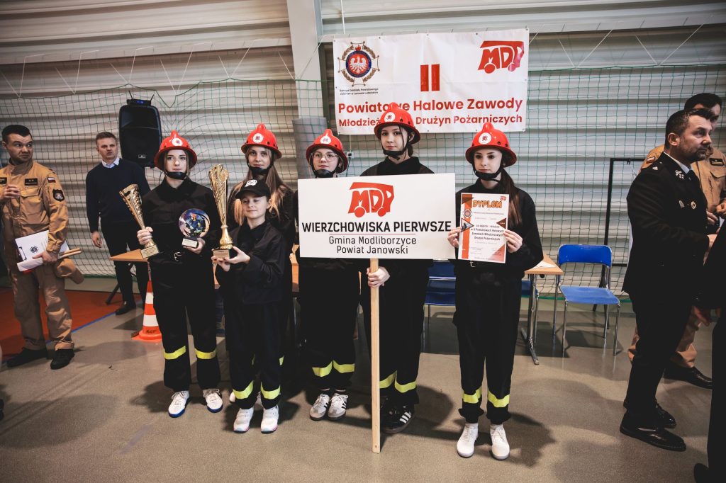 Grupa młodych strażaczek w czarnych mundurach i czerwonych hełmach stoi na hali sportowej, trzymając puchary, dyplom i tablicę z napisem Wierzchowiska Pierwsze.