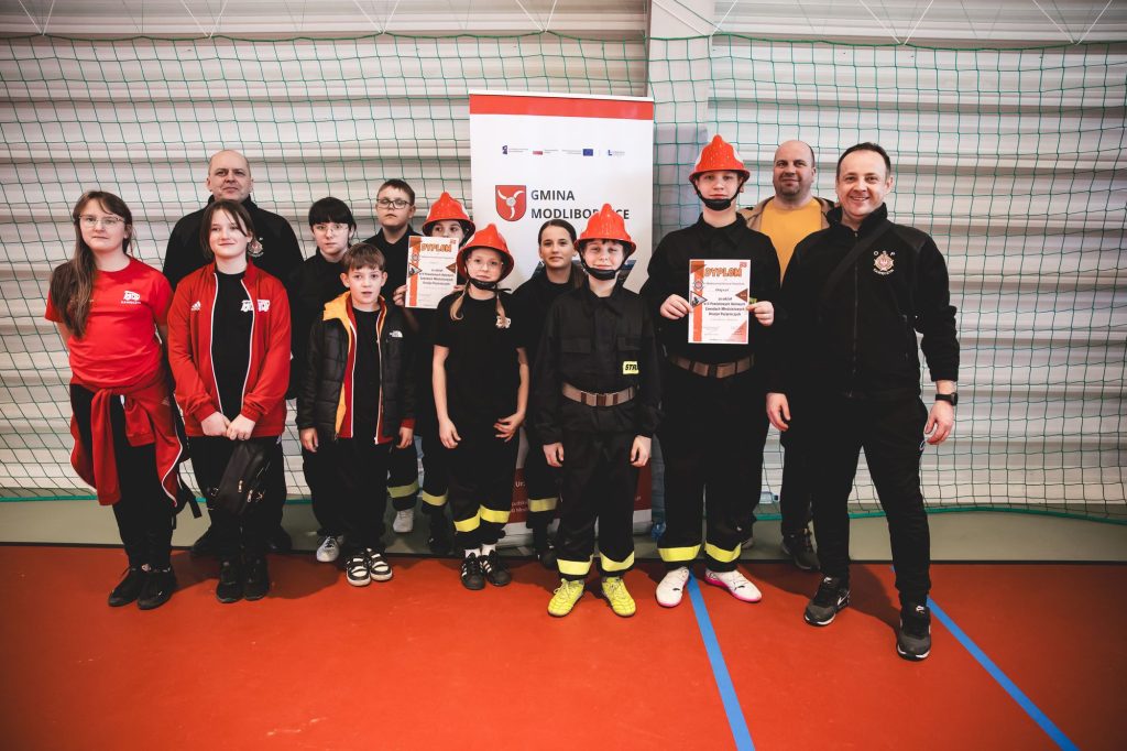 Grupa dzieci i dorosłych na hali sportowej, dzieci w czarnych strojach strażackich z czerwonymi hełmami, dwie osoby trzymają dyplomy, tło z siatką i banerem.