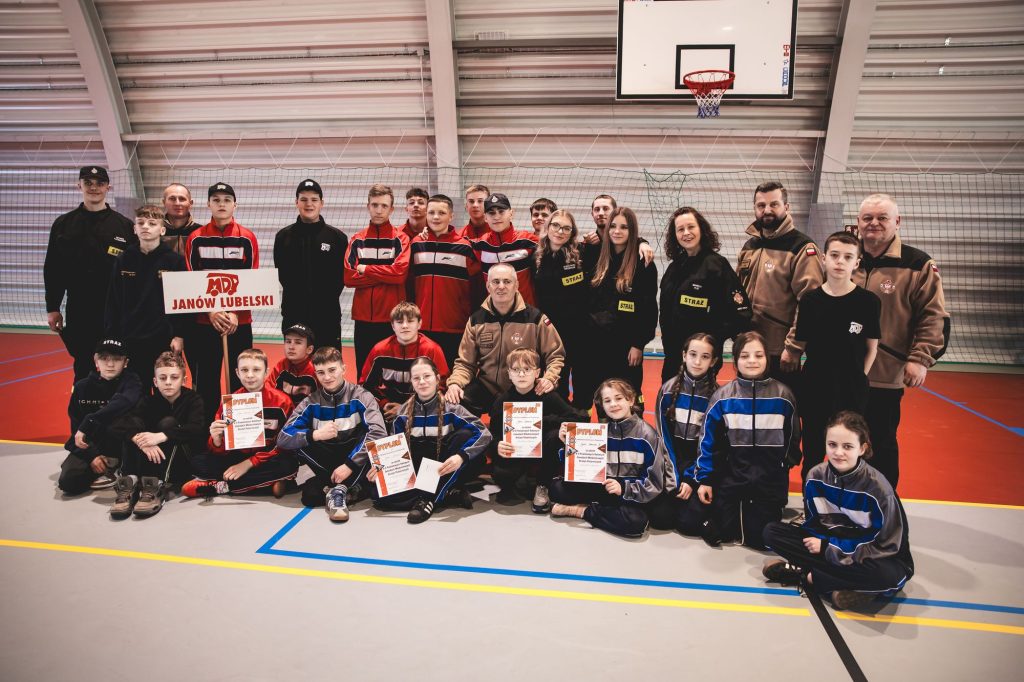 Grupa dzieci i dorosłych w sportowych strojach, część trzyma dyplomy i tabliczkę "MDP Janów Lubelski", na tle sali gimnastycznej z koszem i siatką.