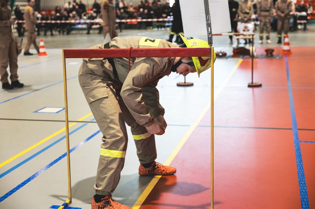 Osoba w beżowym kombinezonie i żółtym kasku ugina się pod niską, czerwoną poprzeczką na hali sportowej z kolorową podłogą.