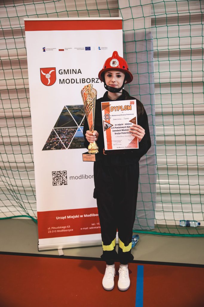Młoda dziewczyna w czarnym stroju strażackim i czerwonym hełmie stoi na hali sportowej, trzyma złoty puchar i dyplom na tle baneru Gmina Modliborzyce.