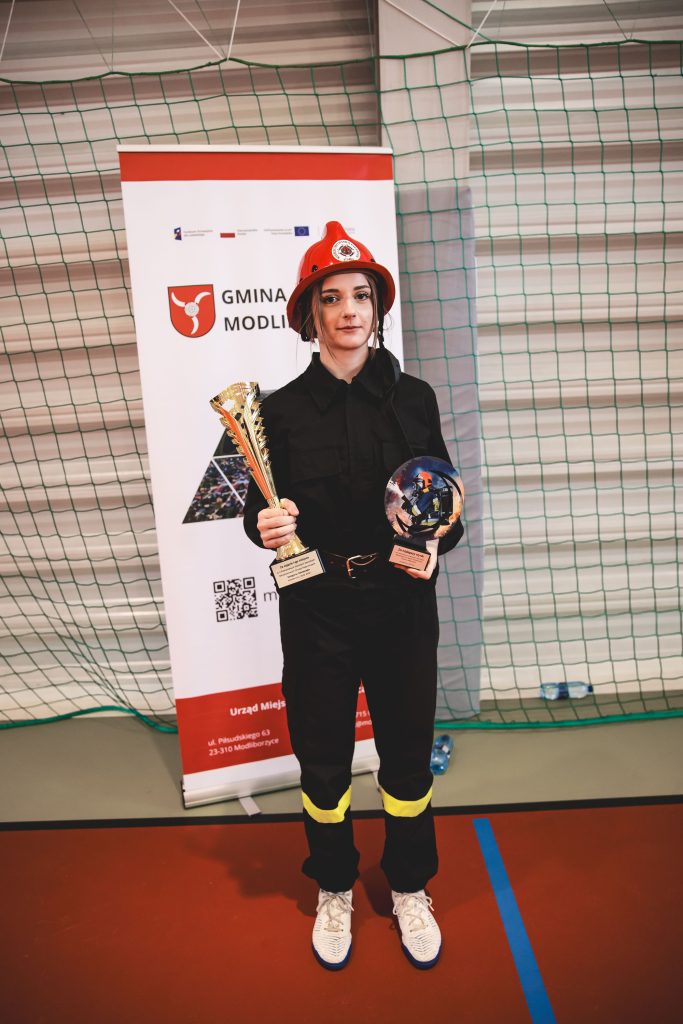 Kobieta w czarnym ubraniu strażackim i czerwonym hełmie stoi na hali sportowej, trzyma złoty puchar i statuetkę, w tle baner z napisem Gmina Modliborzyce.