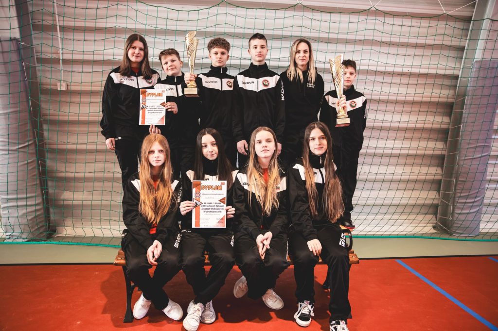 Dziesięcioro młodych osób w czarno-białych dresach sportowych, dwie osoby trzymają złote puchary, dwie dyplomy, stoją i siedzą na ławce na tle zielonej siatki.