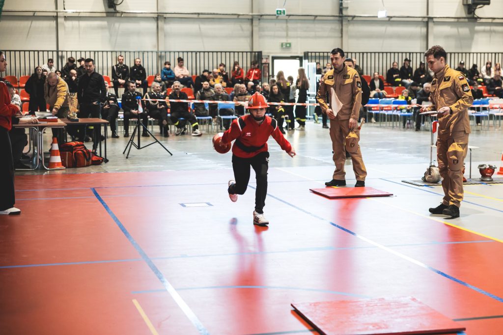 Chłopiec w czerwonym kasku i ubraniu biegnący po czerwonej hali sportowej, obok dwaj mężczyźni w beżowych mundurach, w tle widownia i stoły z sędziami.
