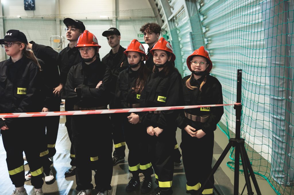 Grupa młodych osób w czarnych mundurach z żółtymi odblaskami i czerwonych hełmach, stojących za biało-czerwoną taśmą wewnątrz hali sportowej z zieloną siatką.
