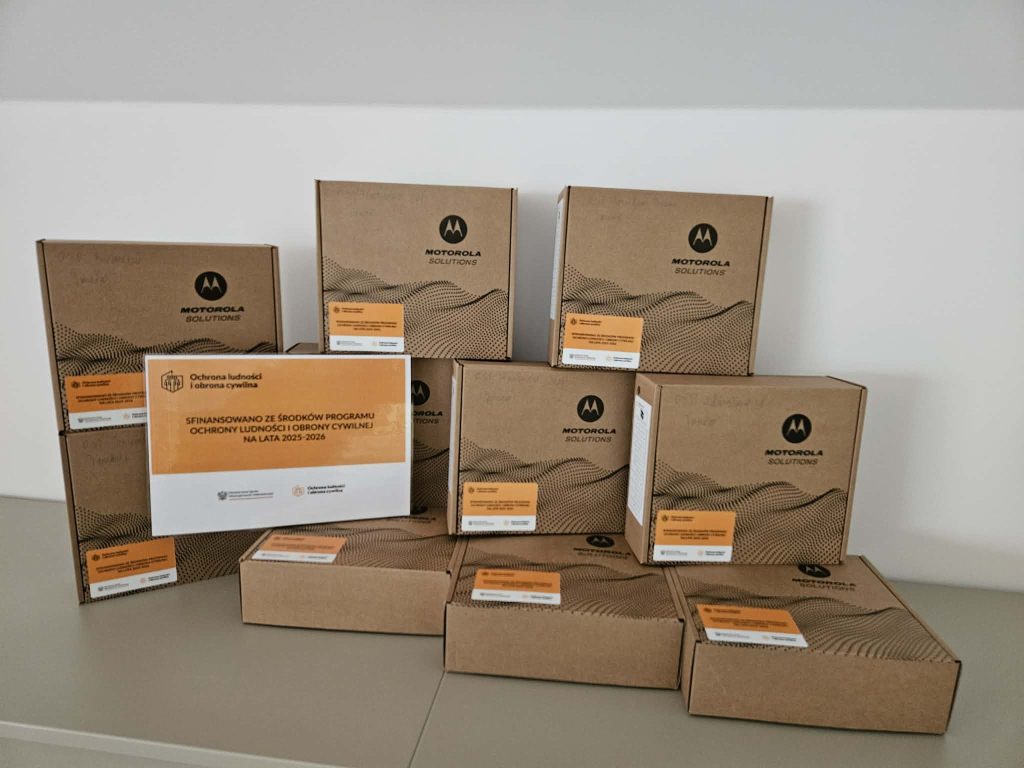 Stosy brązowych kartonów Motorola Solutions z pomarańczowymi naklejkami i białą planszą z napisem o finansowaniu programu ochrony ludności i obrony cywilnej.