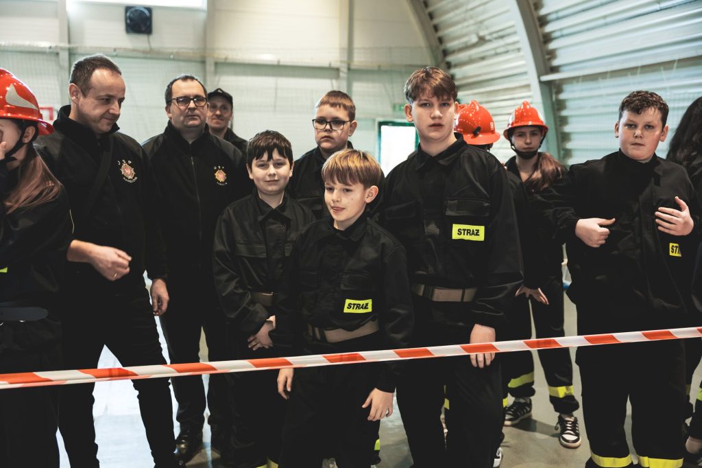 Grupa dzieci i dorosłych w czarnych mundurach strażackich z żółtymi naszywkami "STRAŻ", niektórzy w czerwonych hełmach, stoją za czerwonobiałą taśmą.