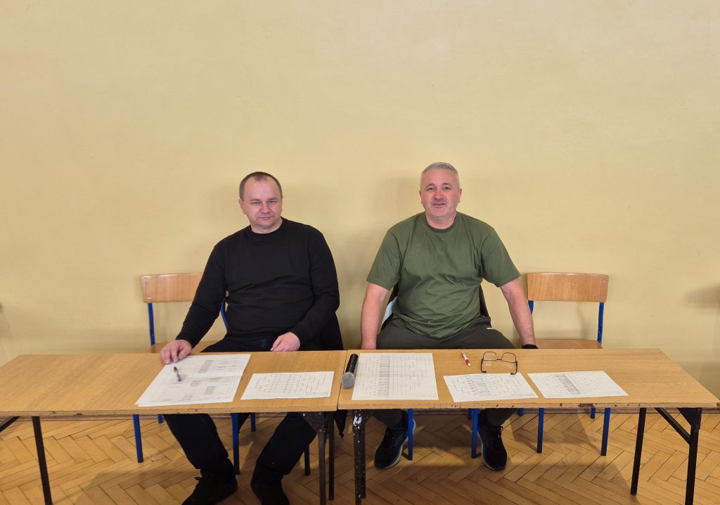 Dwaj mężczyźni siedzą na drewnianych krzesłach za stołem z jasnego drewna, na stole rozłożone kartki papieru i mikrofon, tło beżowa ściana, podłoga drewniana.