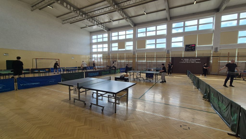 Hala sportowa z parkietową podłogą, stoły do tenisa stołowego, kilka osób grających, duże okna, tablica wyników, ścianka z napisem turniej strażaków.