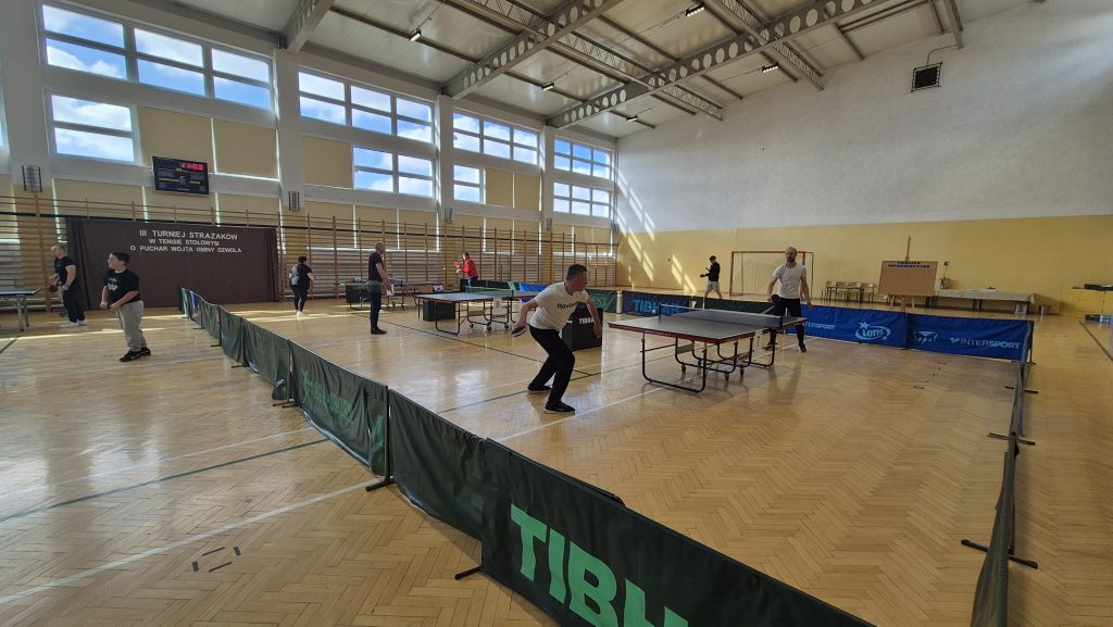 Hala sportowa z dużymi oknami, na parkiecie kilka stołów do tenisa stołowego, ludzie grający w tenisa, drewniana podłoga, barierki z logo TIBHAR i banery reklamowe.