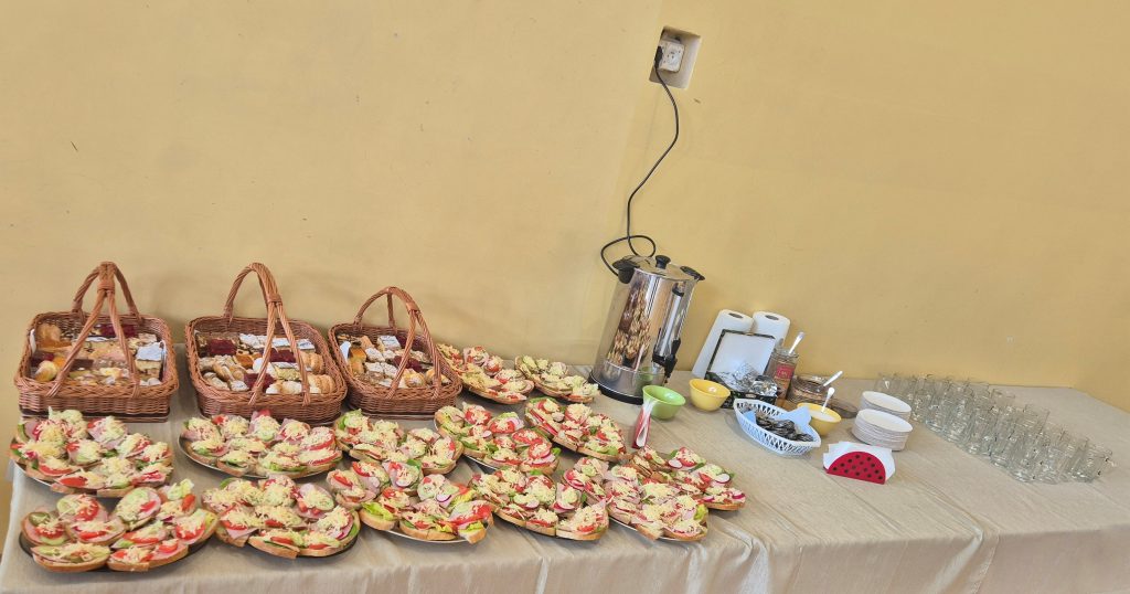 Stół nakryty beżowym obrusem z trzema koszykami pełnymi słodkości, wieloma kanapkami z warzywami i serem, metalowym podgrzewaczem, naczyniami, szklankami i serwetnikiem.