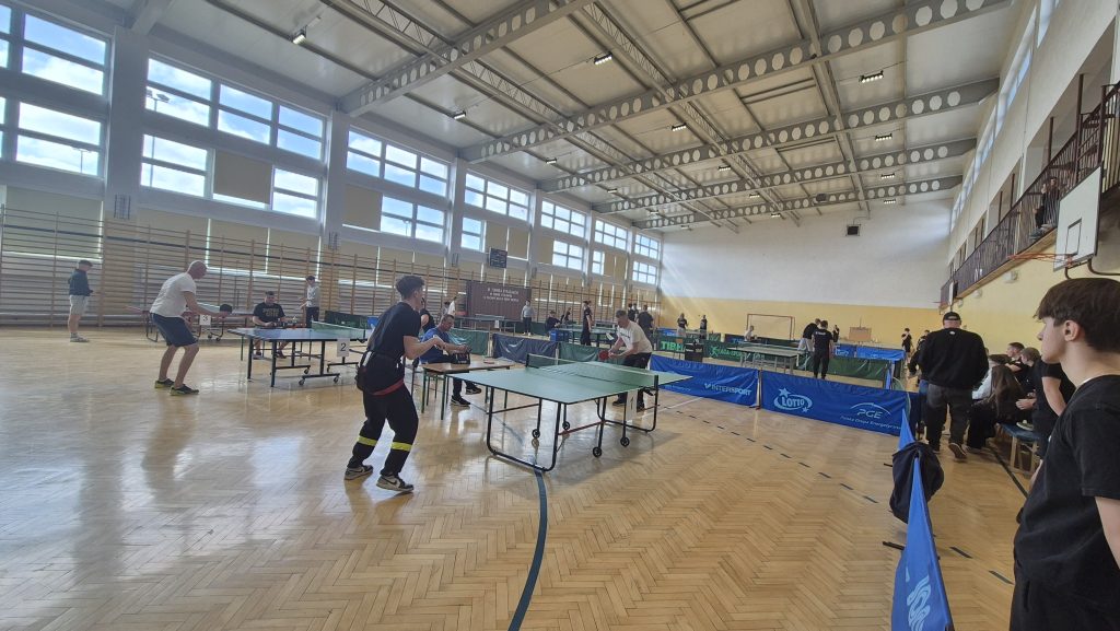 Hala sportowa z dużymi oknami, drewnianą podłogą, stołami do tenisa stołowego, zawodnikami i widzami ubranymi głównie na czarno.